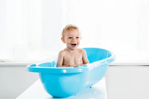 Cheerful toddler kid smiling while taking bath in blue baby bathtub — Stock Photo