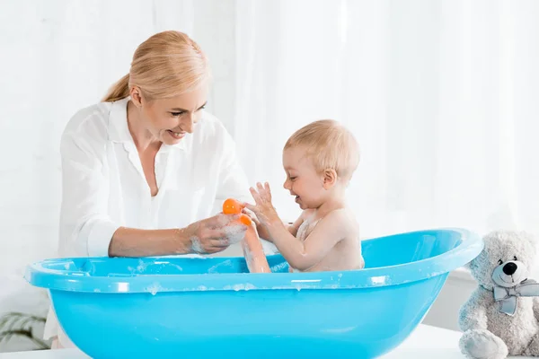 Enfant tout-petit heureux près de mère blonde prenant un bain à la maison — Photo de stock