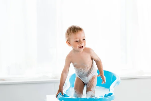 Niño feliz sonriendo mientras está de pie en la bañera azul del bebé - foto de stock
