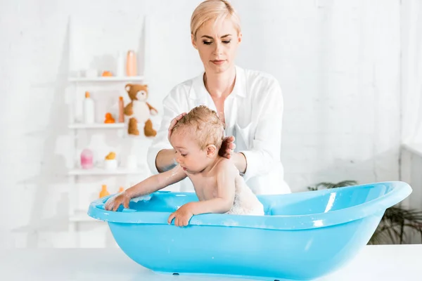 Attractive blonde mother touching hair of cute toddler son in bathroom — Stock Photo