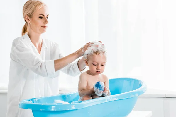 Atractiva madre lavando cabeza de lindo hijo pequeño en el baño - foto de stock
