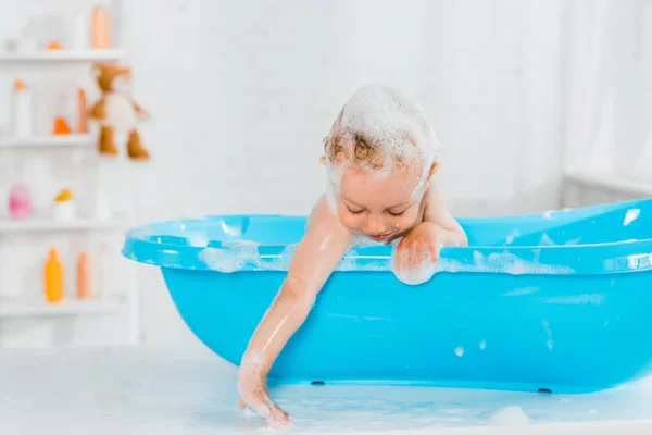 Lindo niño sonriendo mientras toca la espuma del baño y tomando baño en la bañera de plástico del bebé - foto de stock