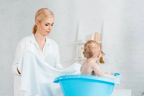 Belle mère blonde tenant serviette près de tout-petit fils dans la salle de bain — Photo de stock