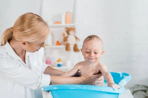 Attractive blonde mother looking at happy toddler son in bathroom — Stock Photo