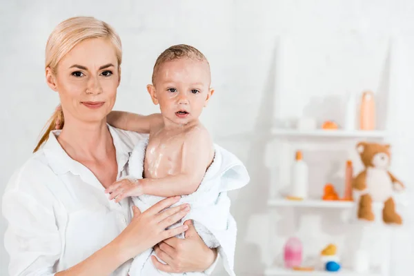 Attractive blonde mother holding in arms wet toddler son in bathroom — Stock Photo