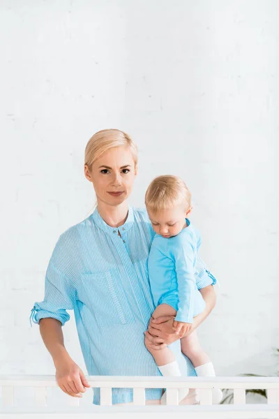 Mãe atraente segurando nos braços filho da criança e de pé perto berço do bebê em casa — Fotografia de Stock