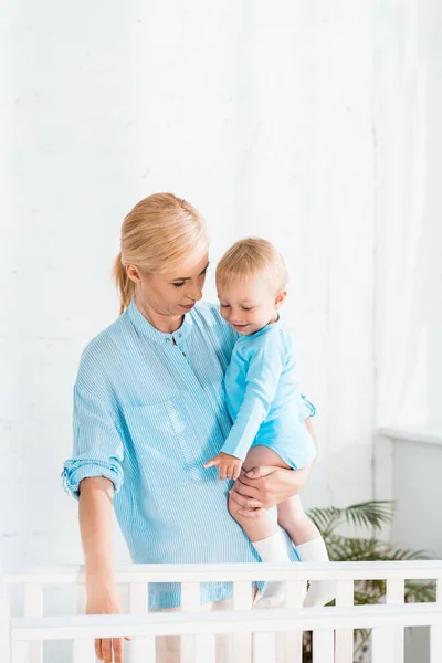 Blonde mother holding toddler son and standing near baby crib at home — Stock Photo