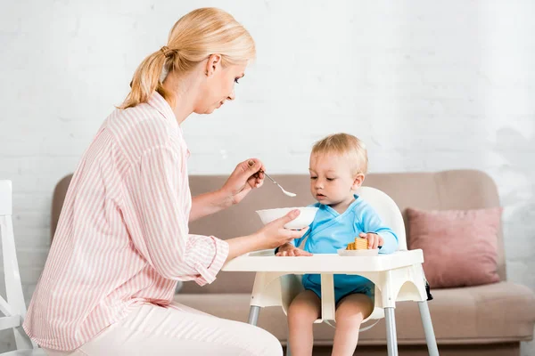 Rubia madre celebración bowl y alimentación lindo niño hijo en casa - foto de stock