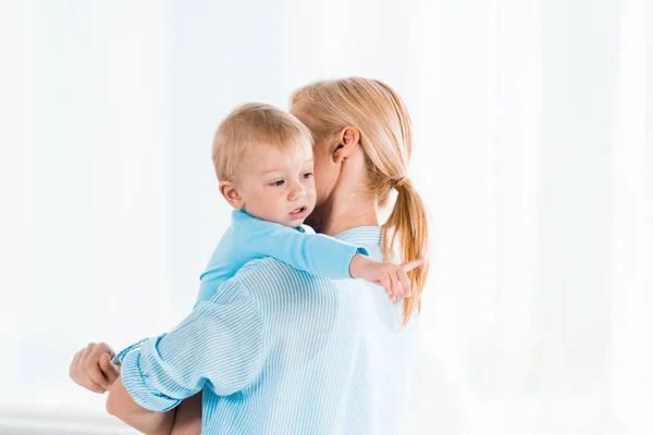 Vue arrière de la femme blonde tenant dans les bras mignon tout-petit fils pointant du doigt à la maison — Photo de stock