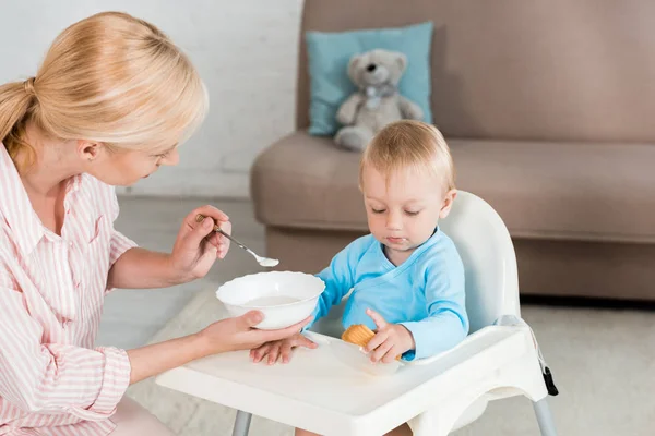Rubia madre celebración bowl y alimentación niño pequeño hijo en casa - foto de stock
