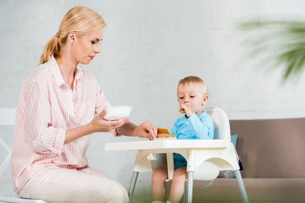 Selective focus of blonde mother holding bowl near cute toddler son in feeding chair — Stock Photo