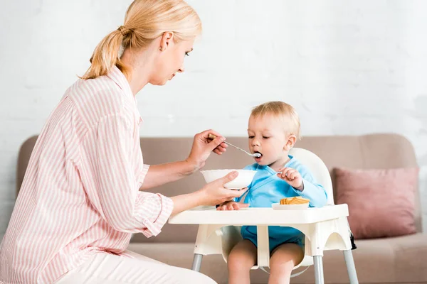 Attraktive blonde Mutter hält Schüssel und füttert niedlichen Kleinkind-Sohn zu Hause — Stockfoto