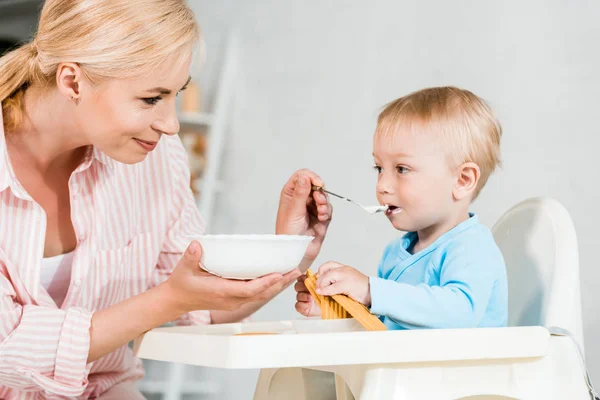 Allegra madre bionda che tiene la ciotola e alimenta il figlio bambino carino a casa — Foto stock