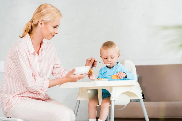 Selektiver Fokus der glücklichen blonden Mutter, die Schale in der Hand hält und ihren niedlichen Kleinkindsohn zu Hause füttert — Stockfoto