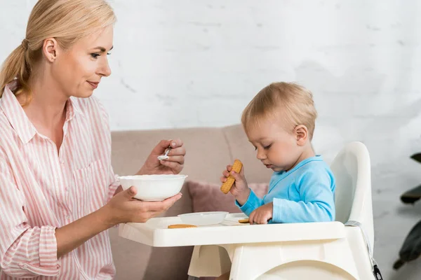 Feliz madre sosteniendo cuchara con comida para bebés cerca de lindo hijo pequeño sentado en silla de alimentación - foto de stock