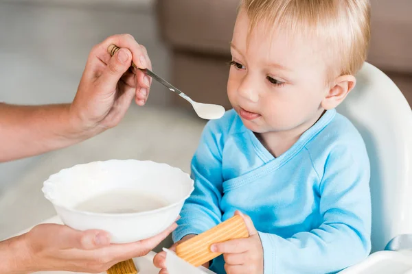 Vista ritagliata della madre che tiene il cucchiaio con il cibo per bambini vicino al figlio bambino carino seduto sulla sedia di alimentazione — Foto stock