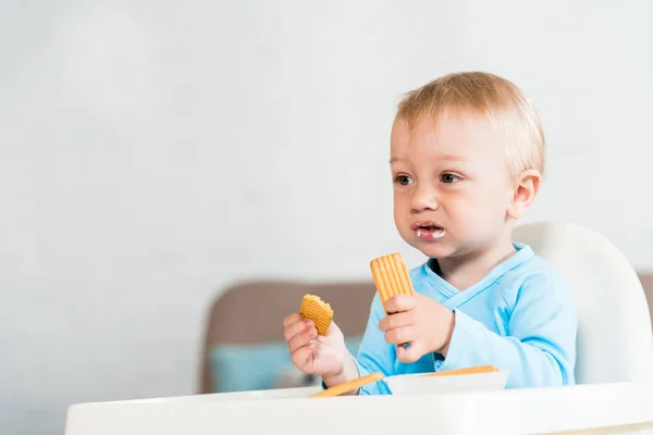 Милый ребенок малыш сидит в кресле для кормления и держит вкусное печенье дома — стоковое фото