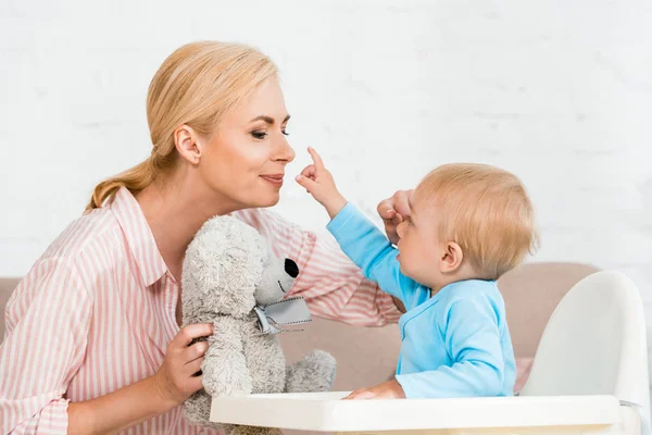 Cute toddler son pointing with finger at cheerful mother with teddy bear — Stock Photo