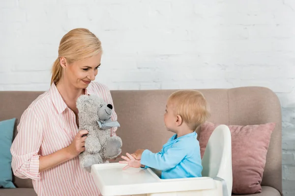 Cheerful mother holding teddy bear near cute toddler son in feeding chir — Stock Photo