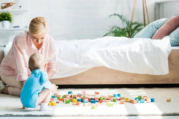 Atractiva madre rubia sentada cerca de hijo pequeño y bloques de juguete en el dormitorio - foto de stock
