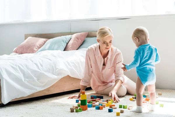 Mère blonde assise sur le tapis près de tout-petit fils jouer avec des briques de construction — Photo de stock