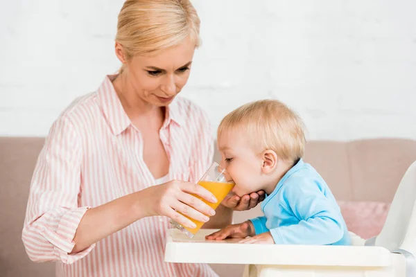 Attractive blonde mother holding glass while toddler son drinking orange juice — Stock Photo