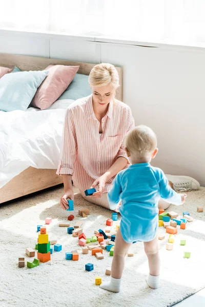 Приваблива блондинка мати сидить на килимі біля дитячого сина, граючи з будівельними цеглами — стокове фото