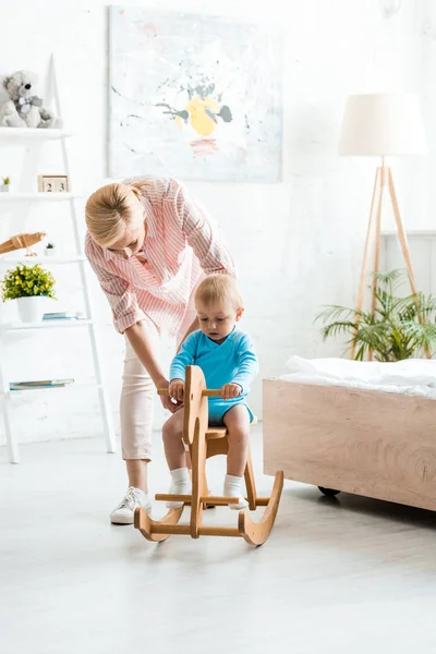 Blonde mother standing near cute toddler son swinging on rocking horse — Stock Photo