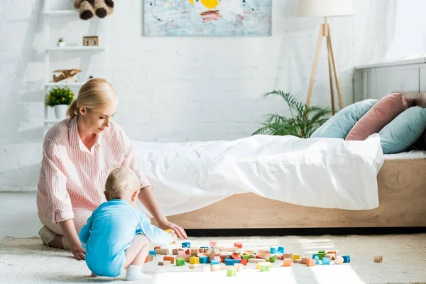 Mujer rubia sentada en la alfombra cerca de hijo pequeño jugando con ladrillos de construcción - foto de stock