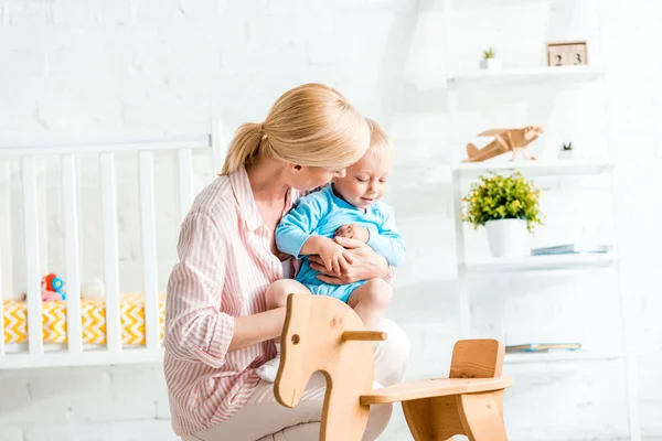 Blonde Mutter hält süßen Kleinkind-Sohn in der Nähe von Schaukelpferd im Arm — Stockfoto