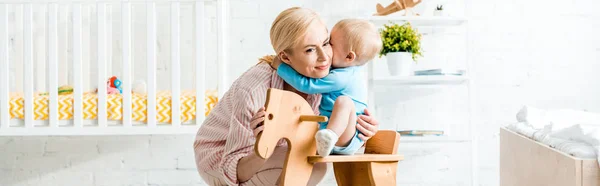 Panoramic shot of cute toddler son hugging happy mother near rocking horse — Stock Photo