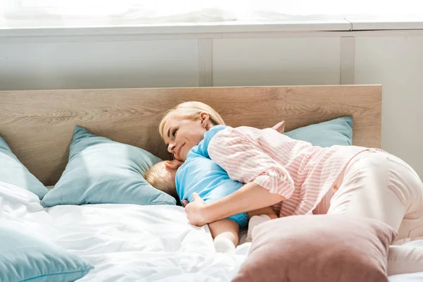Feliz madre abrazando hijo pequeño mientras está acostado en la cama y abrazando hijo pequeño - foto de stock