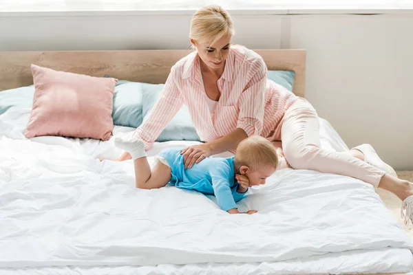Cheerful blonde mother looking at cute toddler son crawling on bed — Stock Photo