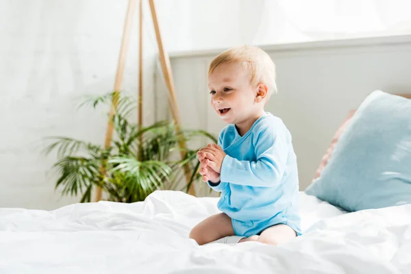 Niño pequeño lindo y sonriente sentado en la cama con las manos apretadas - foto de stock