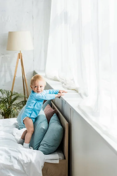 Милий і усміхнений дитина стоїть на ліжку в сучасній спальні — стокове фото
