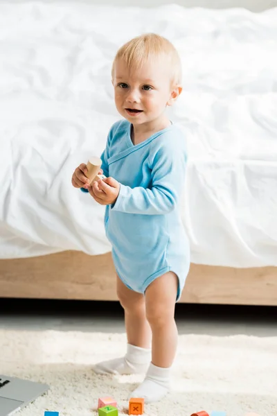 Smiling toddler kid standing with wooden toy near bed — Stock Photo