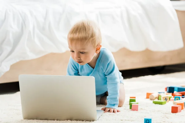 Carino bambino bambino guardando computer portatile vicino blocchi di giocattoli colorati su tappeto — Foto stock