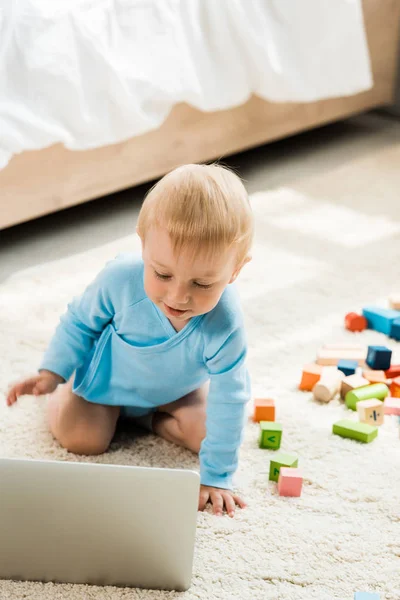 Selective focus of happy toddler kid looking at laptop near colorful toy blocks on carpet — Stock Photo