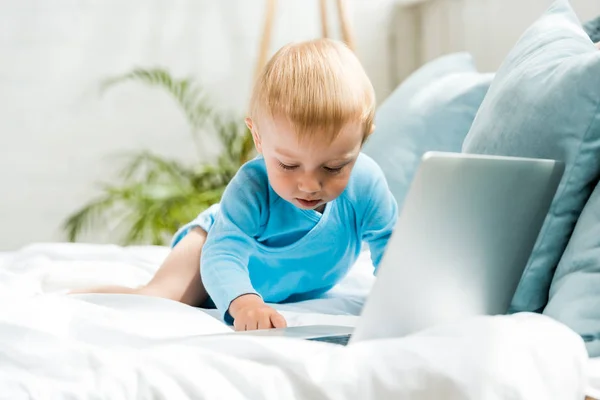 Selective focus of cute toddler kid sitting on bed near laptop at home — Stock Photo