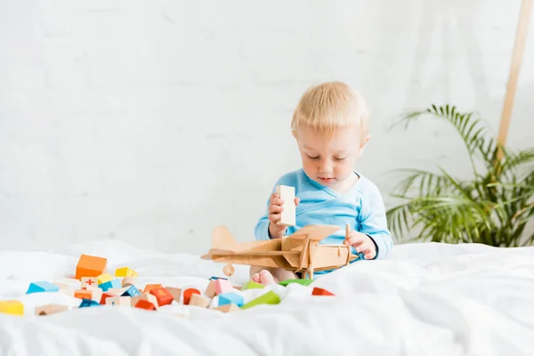 Cute toddler boy playing with wooden biplane near colorful toy blocks on bed — Stock Photo