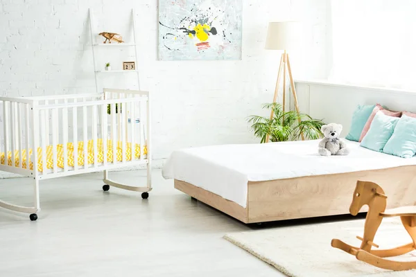 Кроватка возле кровати с белыми постельными принадлежностями и подушками рядом с плюшевым мишкой и лошадью-качалкой — стоковое фото