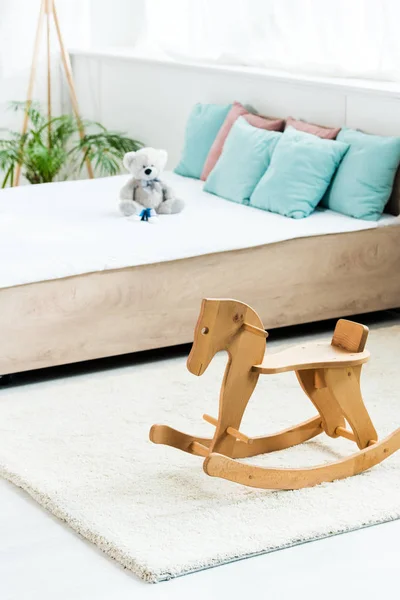 Selective focus of wooden rocking horse near bed with white bedding, pillows and teddy bear — Stock Photo