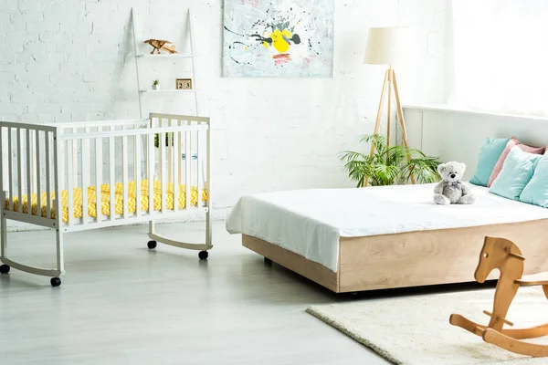 Baby crib near bed with white bedding and pillows near teddy bear and wooden rocking horse — Stock Photo