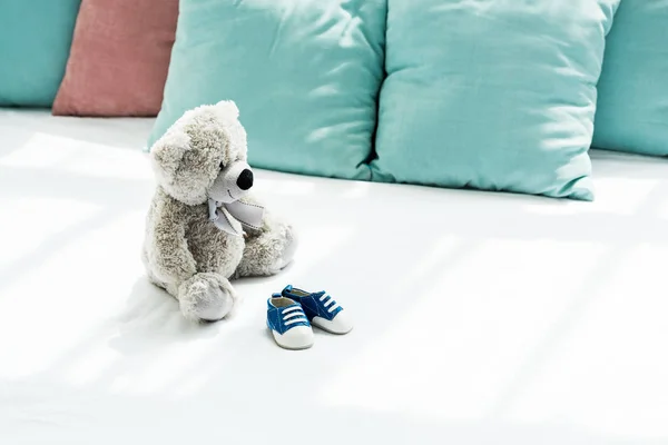 Grey teddy bear near baby sneakers and pillows on white — Stock Photo