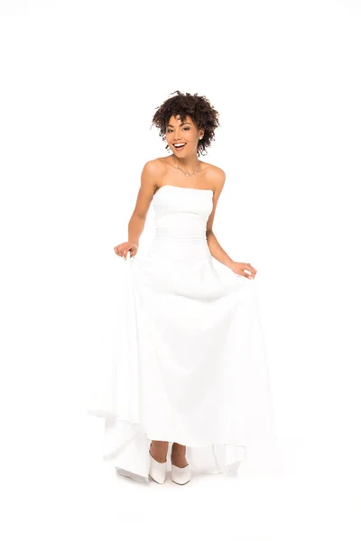 Heureuse mariée afro-américaine souriante tout en touchant robe de mariée isolé sur blanc — Photo de stock