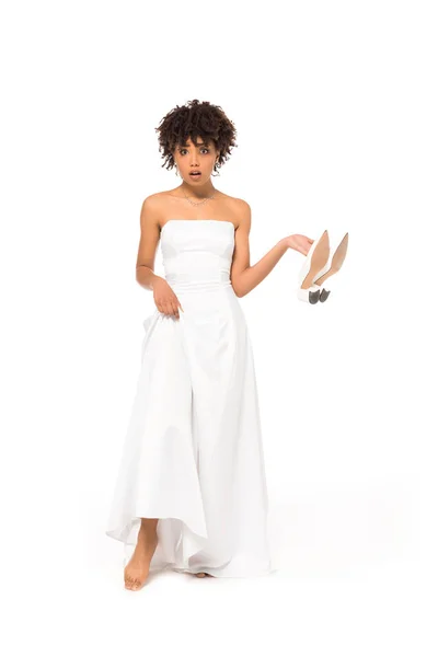 Surpris mariée afro-américaine touchant robe de mariée et tenant des chaussures tout en se tenant isolé sur blanc — Photo de stock