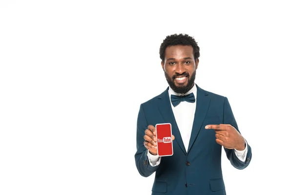 KYIV, UKRAINE - MAY 16, 2019: Happy african american bridegroom pointing with finger at smartphone with youtube app on screen isolated on white — Stock Photo