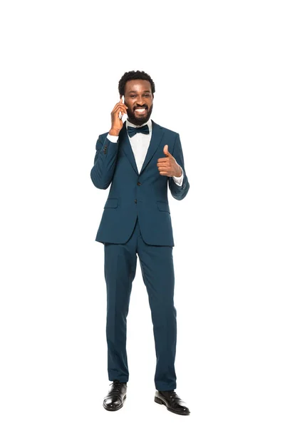 Happy african american man in suit talking on smartphone and showing thumb up isolated on white — Stock Photo