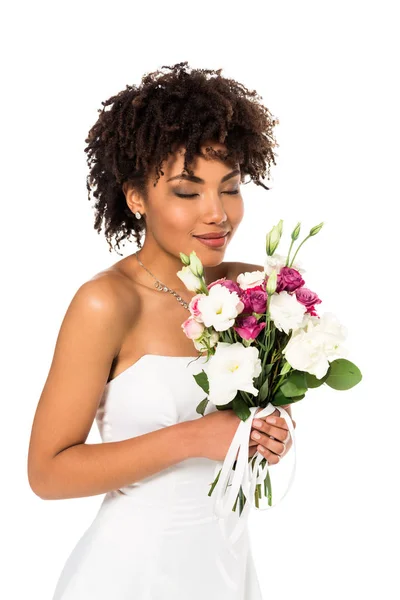 Happy african american bride holding bouquet while smelling flowers isolated on white — Stock Photo