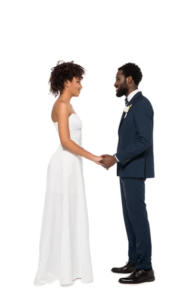 Felice sposo afroamericano e sposa che si tengono per mano mentre si guardano isolati su bianco — Foto stock